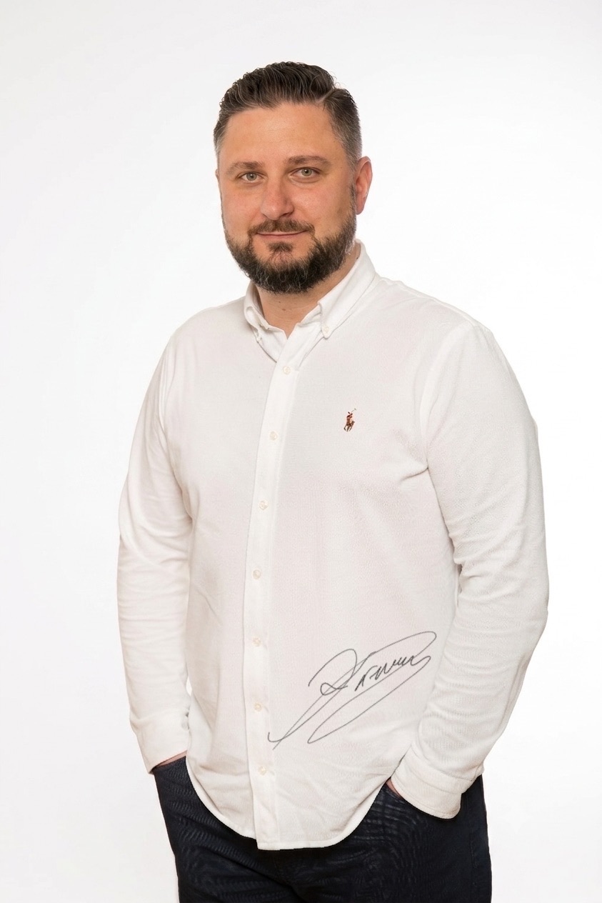 Muž v bílé košili s výšivkou a tmavými džíny, čelně pózuje na světlém pozadí.